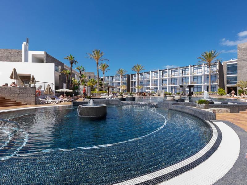 Piscine d'hôtel moderne avec terrasse ensoleillée, transats et palmiers sous un ciel dégagé.