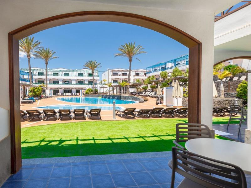 Vue à travers une arche sur une piscine d'hôtel avec des chaises longues et des palmiers sous un ciel clair.