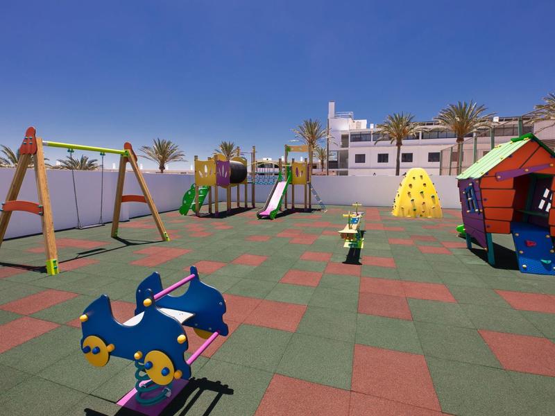 Aire de jeux colorée pour enfants avec balançoires, toboggans et maisonnette sous ciel bleu.