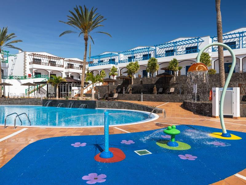 Bassin pour enfants avec jeux d'eau à côté d'une grande piscine et de plusieurs bâtiments aux accents bleus.