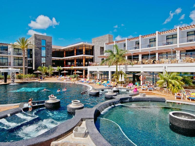 Complex hôtelier moderne avec plusieurs piscines, terrasses ensoleillées et palmiers sous un ciel bleu.