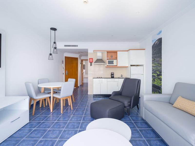 Salon lumineux avec canapé gris, table, chaises et kitchenette dans un appartement moderne.