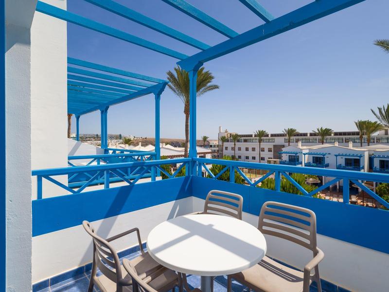 Balcon avec rambarde bleue, table ronde blanche et chaises avec vue sur l'hôtel et les palmiers.
