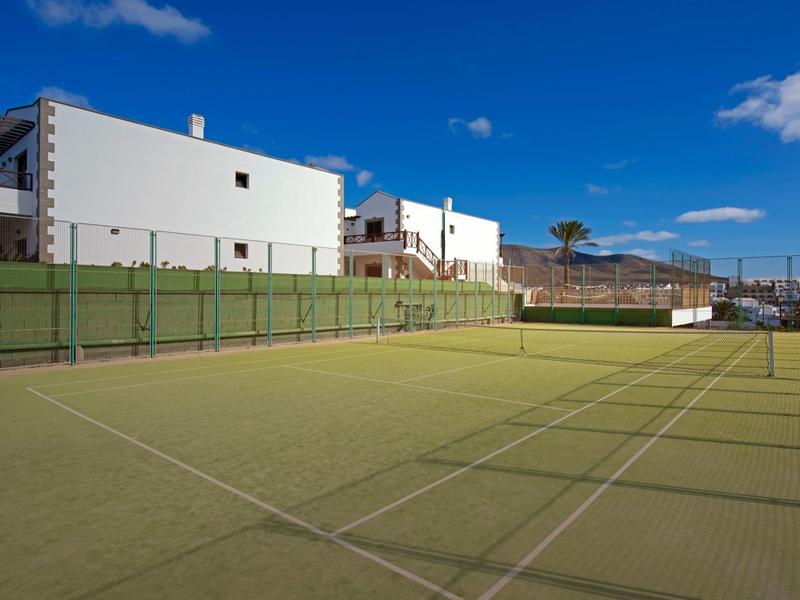 Terrain de tennis vide avec une clôture verte et des bâtiments blancs en arrière-plan sous un ciel bleu.