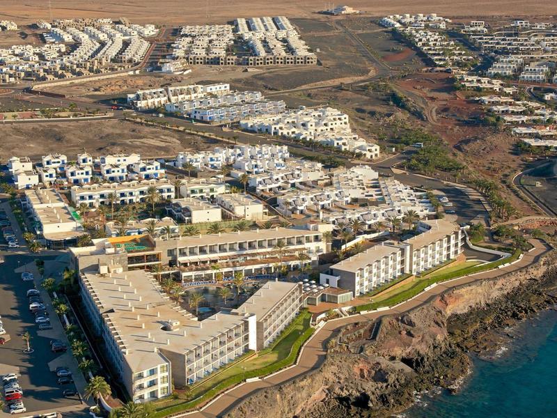 Vue d'un complexe hôtelier avec des bâtiments blancs sur une côte rocheuse et des montagnes en arrière-plan.