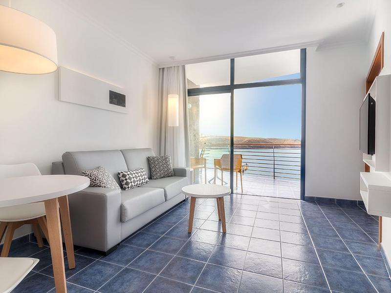 Salon moderne avec canapé gris, petite table et accès au balcon avec vue sur la mer.