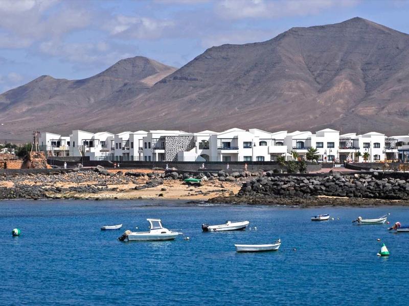 White holiday homes on the shore with small boats in clear blue water and mountains in the background.