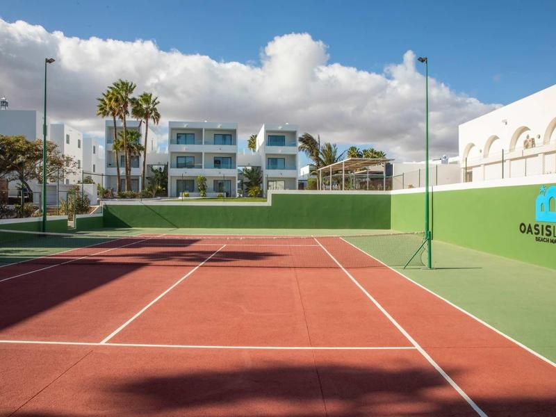 Red tennis court with white lines in front of modern white buildings and palm trees under a blue sky.