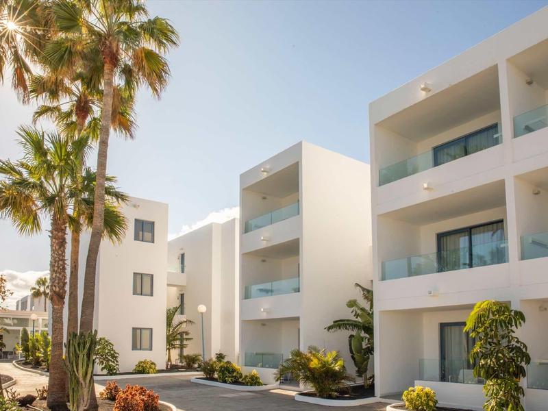 Modern white hotel buildings with balconies and palm trees under sunny weather.