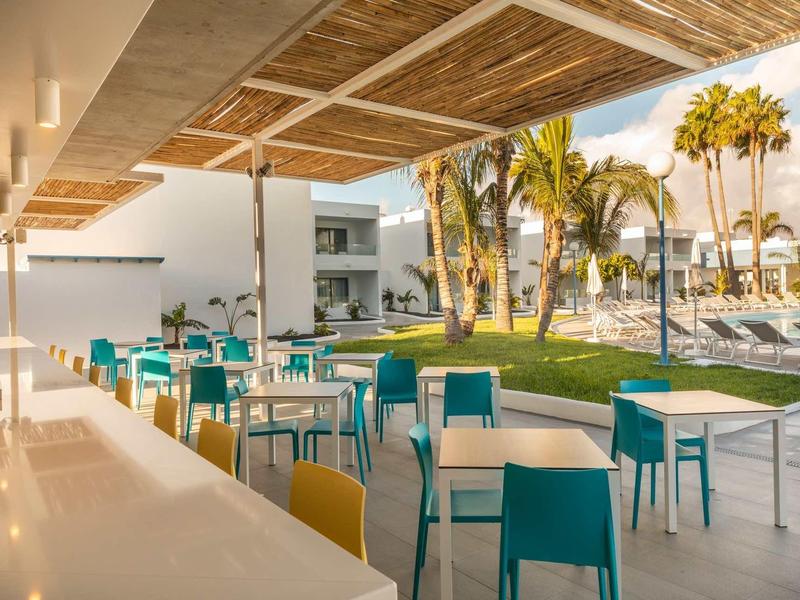 Modern outdoor area with colorful chairs and tables beside a hotel pool and palm trees.