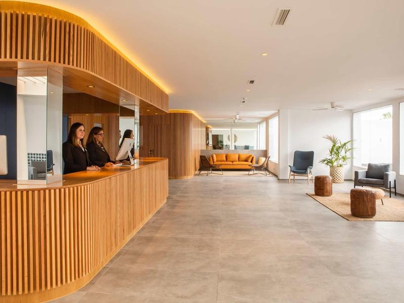 Bright, modern hotel lobby with wooden reception desk, seating areas, and large windows.
