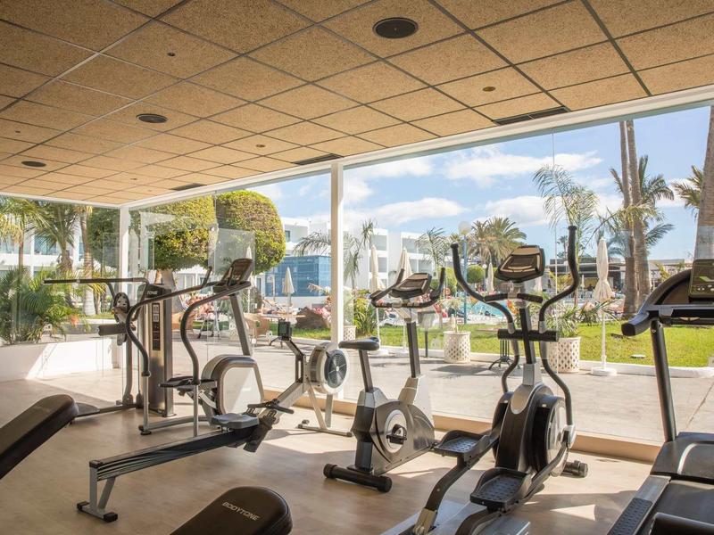 Bright gym with modern equipment and large glass windows overlooking garden and pool.