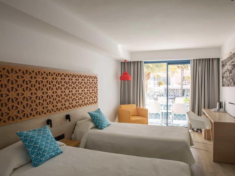 Hotel room with two single beds, blue patterned pillows, orange armchair, and balcony with sea view.
