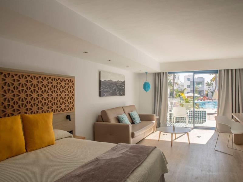 Bright hotel room with double bed, sofa, chairs, and balcony overlooking pool and outdoor area.