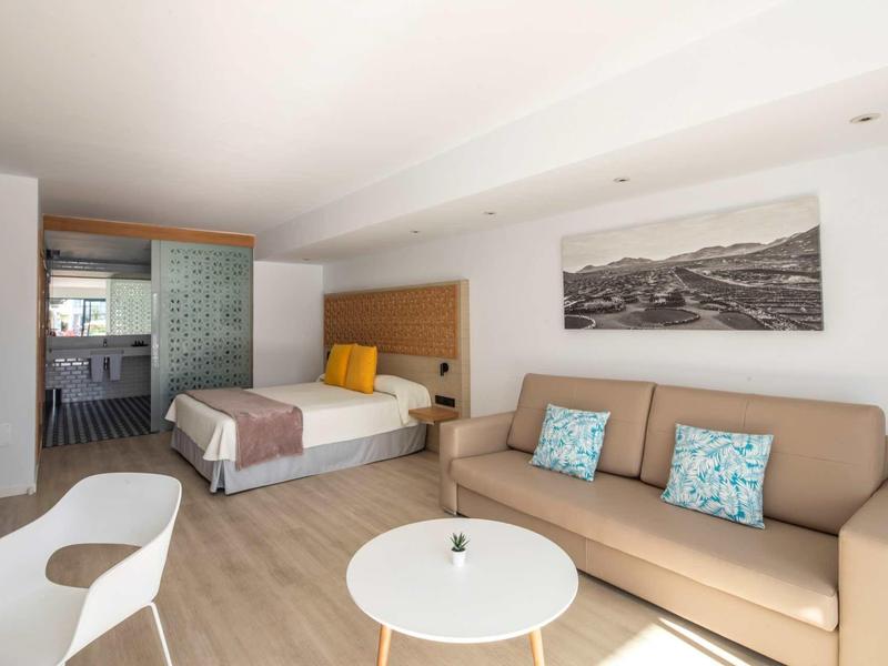 Modern hotel room with double bed, sofa, round table, and kitchenette in the background.