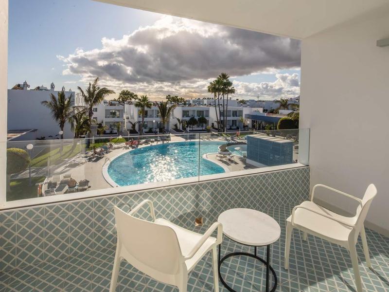Balcony with table and two chairs overlooking pool and hotel buildings under cloudy sky.