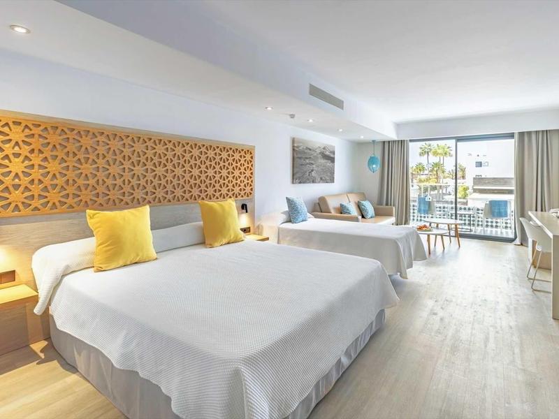 Bright hotel room with two double beds, yellow pillows, and balcony with sea view.