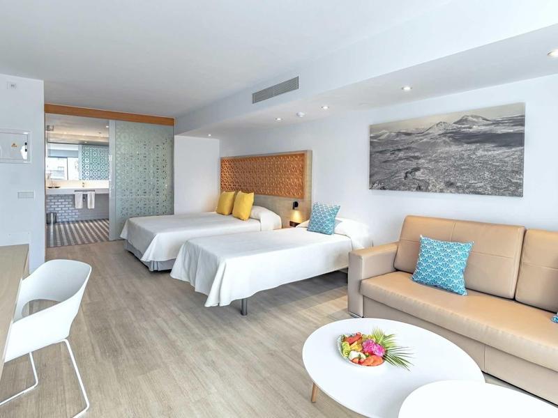 Bright hotel room with two double beds, sofa, side table, and modern decor.