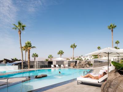 Moderner Poolbereich mit Liegen, Sonnenschirmen und Palmen unter klarem blauem Himmel.