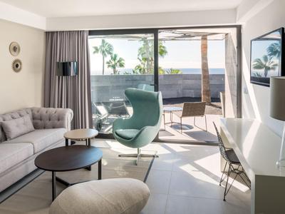 Modern eingerichtetes Wohnzimmer mit Sofa, Sesseln und großer Glasfront zum Balkon mit Palmenblick.