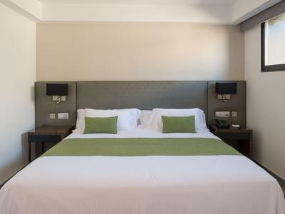 Schlichtes Schlafzimmer mit großem Bett, weißen Laken, grünen Kissen und zwei Nachttischen aus Holz.