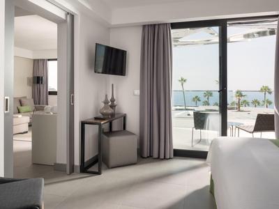 Modern eingerichtetes Hotelzimmer mit großem Fenster, Blick auf Terrasse und Meer, hellen Vorhängen.