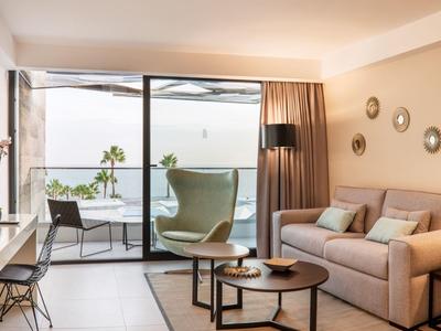 Helles Wohnzimmer mit Sofa, Sessel, Tisch, Lampen und großem Fenster mit Blick auf Meer und Balkon.