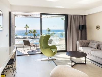 Heller Wohnraum mit grauem Sofa, grünem Sessel, großem Fenster und Meerblick mit Palmen.