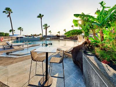 Terrasse mit Stühlen, Tischen, Palmen und einem Pool bei strahlendem Sonnenschein.