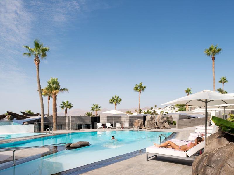 Moderner Poolbereich mit Liegen, Sonnenschirmen und hohen Palmen unter klarem blauem Himmel.