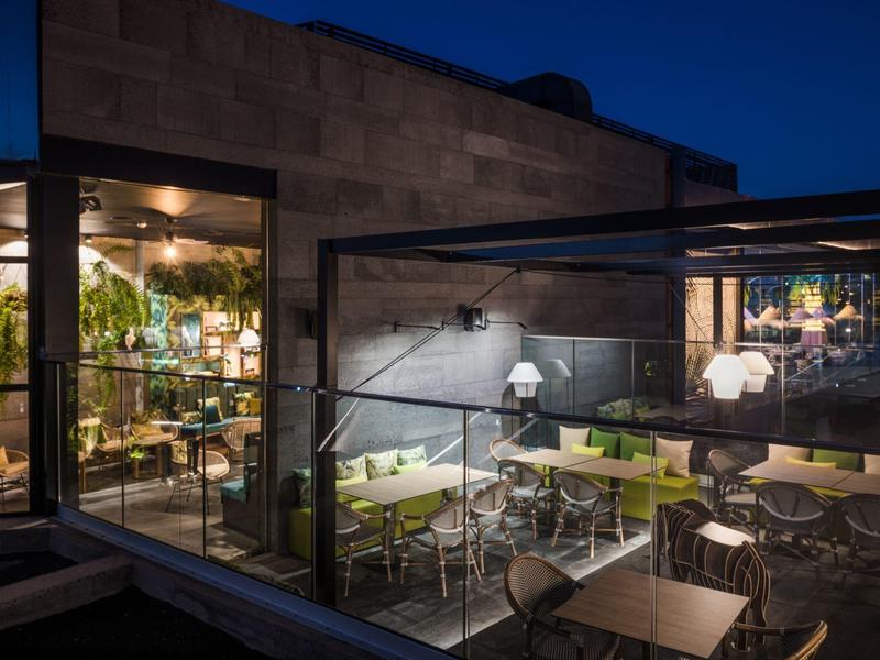 Moderne Außenterrasse eines Cafés mit Tischen, Stühlen und grünen Pflanzen bei Abenddämmerung.