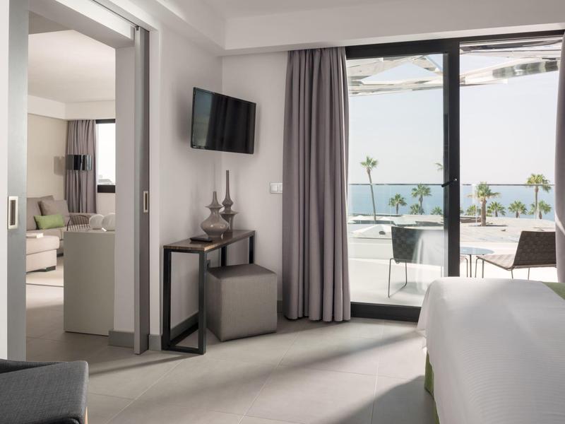 Modern eingerichtetes Hotelzimmer mit großem Fenster, Blick auf Terrasse und Meer, hellen Vorhängen.