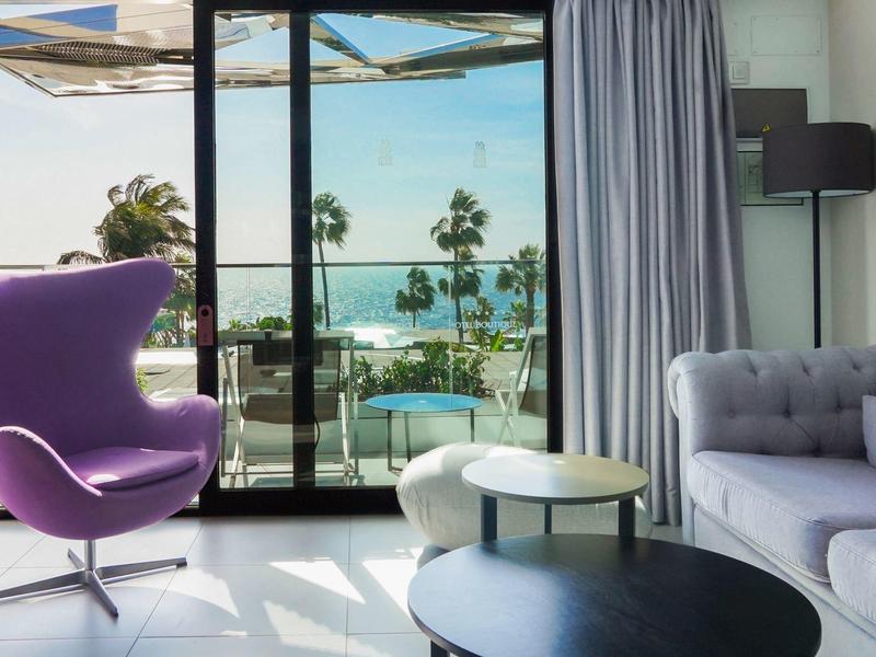 Modernes Wohnzimmer mit lila Sessel, grauem Sofa, Couchtischen und Blick auf Terrasse mit Palmen.