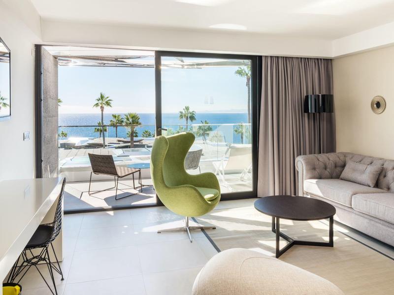 Heller Wohnraum mit grauem Sofa, grünem Sessel, großem Fenster und Meerblick mit Palmen.