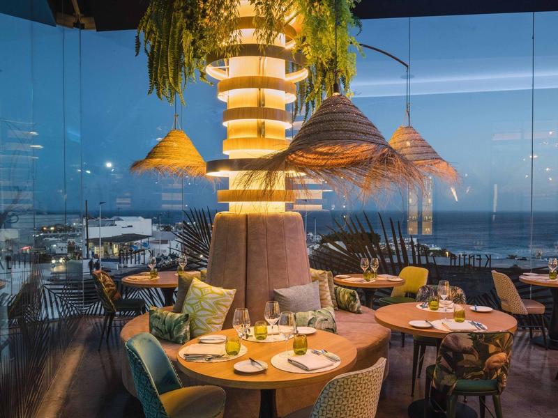 Moderne Bar mit runden Tischen, bunten Stühlen und großen Hängelampen bei Abenddämmerung.