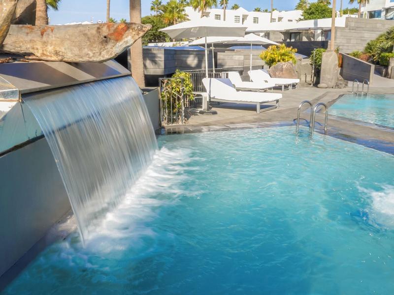 Pool mit Wasserfall, bläulichem Wasser, Sonnenliegen und modernen Architektur im Hintergrund.