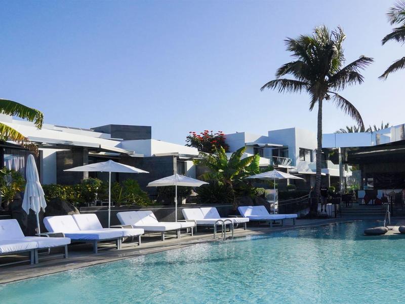 Moderner Poolbereich mit Liegestühlen, Sonnenschirmen und Palmen unter blauem Himmel.