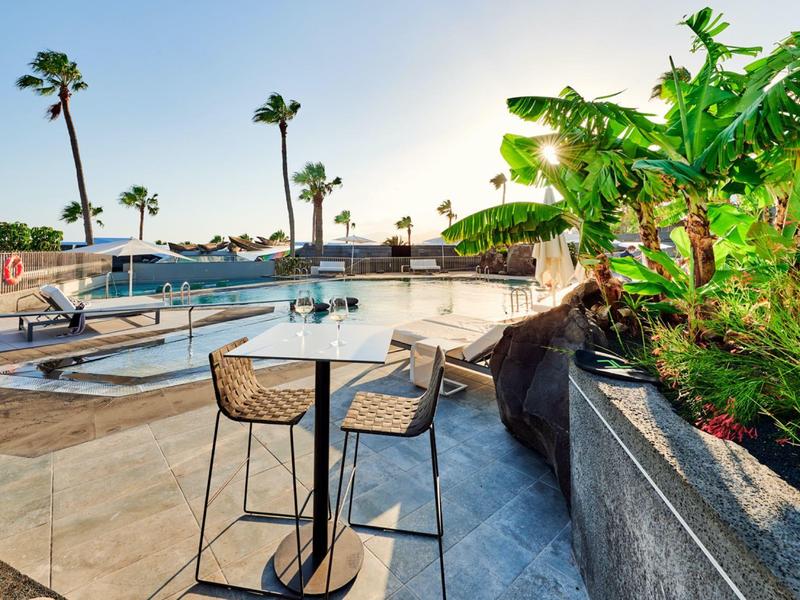 Terrasse mit Stühlen, Tischen, Palmen und einem Pool bei strahlendem Sonnenschein.