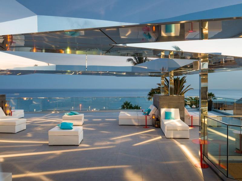 Moderne Terrasse mit weißen Möbeln, Glasgeländer und Meerblick bei sonnigem Wetter.