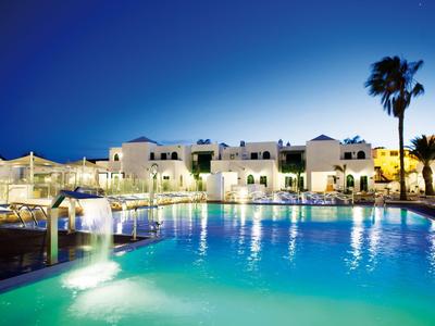 Beleuchteter Hotelpool bei Nacht mit Wasserspiel und Palmen im Hintergrund.