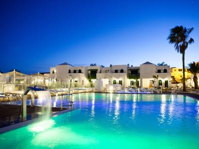 Piscina de hotel iluminada por la noche con cascada y palmeras bajo cielo azul.