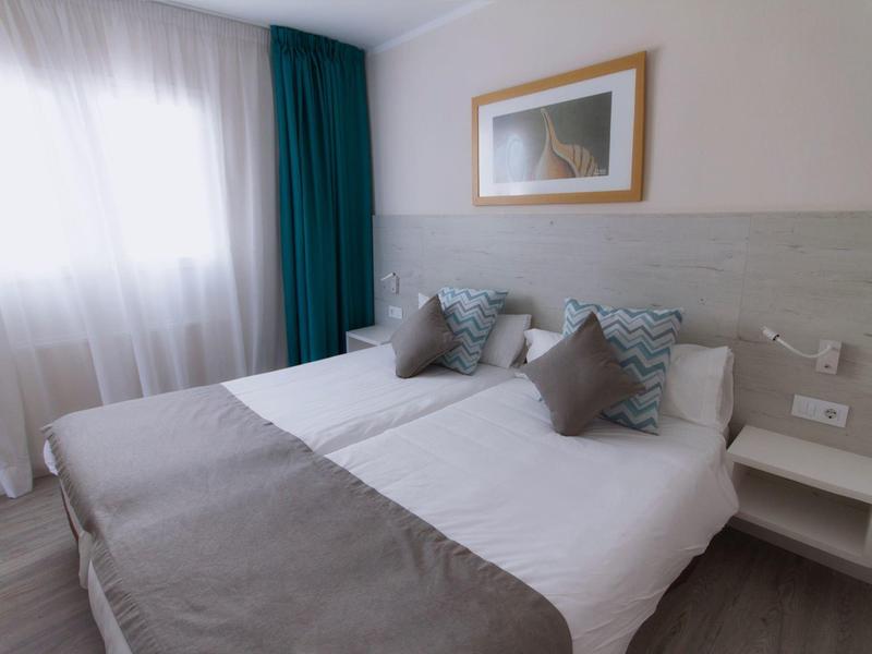 Habitación de hotel luminosa con dos camas individuales, cojines y colcha grises, cortinas y cuadro en la pared.