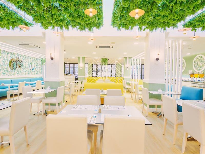 Salón de hotel luminoso con asientos modernos, paredes blancas y techo con plantas verdes colgantes.