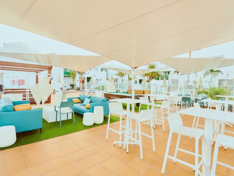 Terraza en la azotea luminosa y moderna con mesas blancas, sillas y sofás azules bajo velas de sombra.