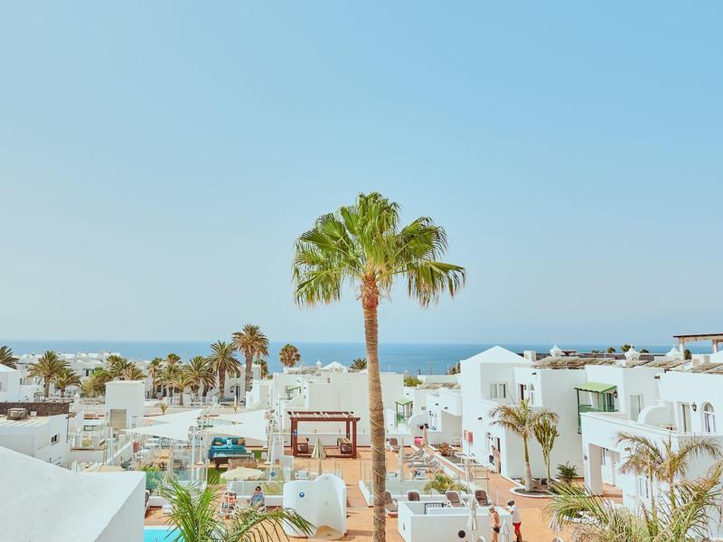 Vista de un lugar vacacional con edificios blancos, palmeras y cielo azul junto al mar.