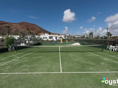 Tennisplatz mit grünem Belag, umgeben von weißen Stühlen, Palmen und Bergen im Hintergrund.