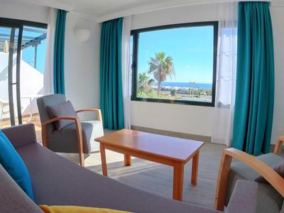 Helles Wohnzimmer mit Sofa, Sesseln, Holztisch, blauen Vorhängen und Meerblick durch große Fenster.