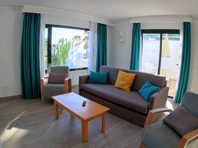 Heller Wohnraum mit Sofa, Sessel, Holztisch, blauen Vorhängen und großen Fenstern mit Außenblick.
