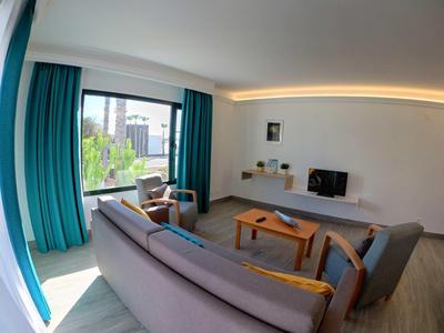 Helles Wohnzimmer mit großen Fenstern, türkisen Vorhängen, Sofa, Sesseln und Holztisch.