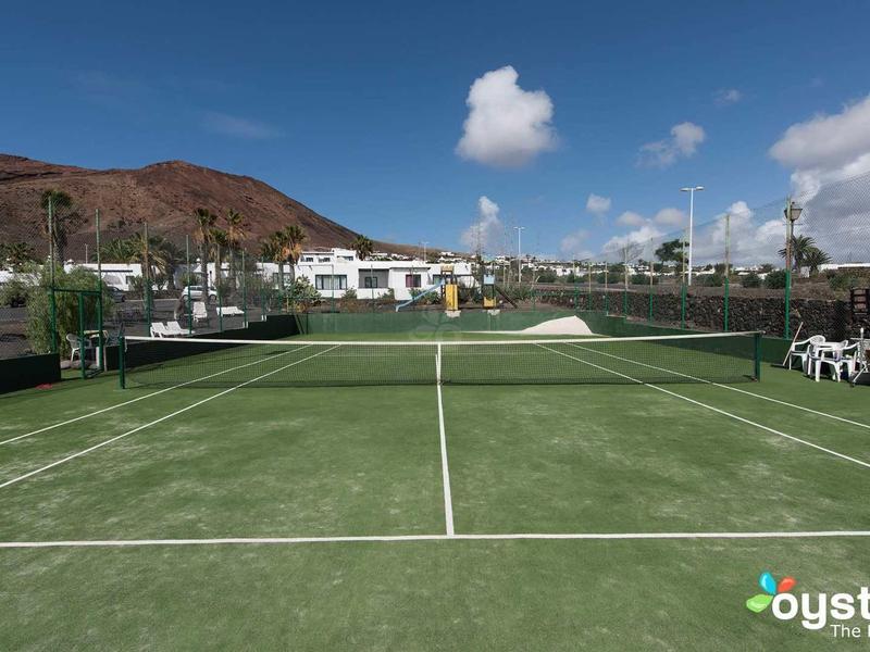Empty tennis court with white lines in front of buildings and hills under a sunny sky.